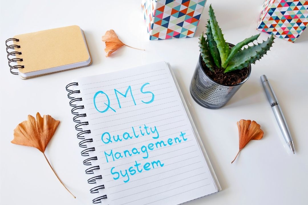 Open notebook with'QMS Quality Management System' written in blue highlighter | Featured image for Is my QMS worth it? Blog by Bramwell Partners.