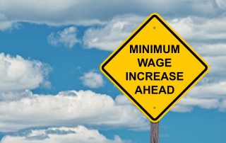 Photo of a yellow diamond traffic sign with the words "minimum wage increase ahead" inside it | Featured image for Minimum Wage Staggered Increase.