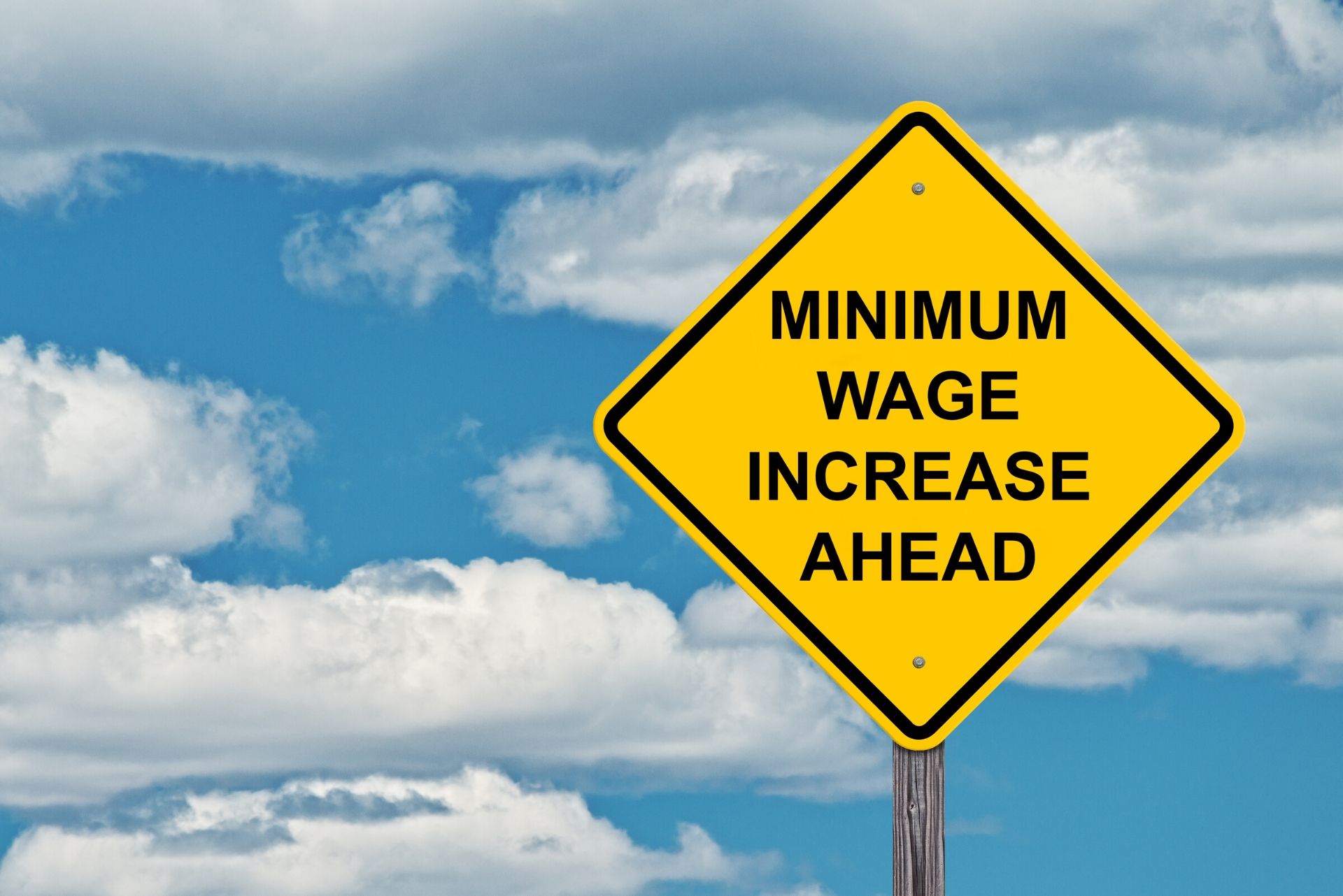 Photo of a yellow diamond traffic sign with the words "minimum wage increase ahead" inside it | Featured image for Minimum Wage Staggered Increase.