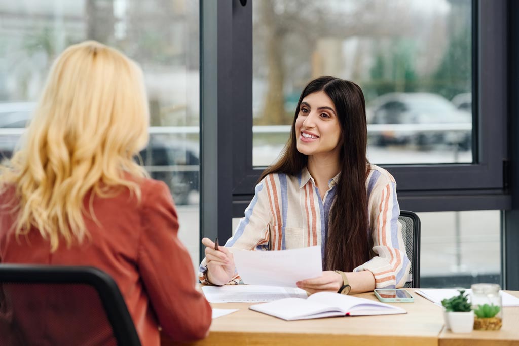 Photo of two women engage in conversation while seated at a desk.